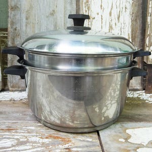 May include: A stainless steel stockpot with a matching lid and black plastic handles on both sides. The pot features a tiered design with a steamer insert, and the metal surface displays engraved text including the brand name Regal Ware.