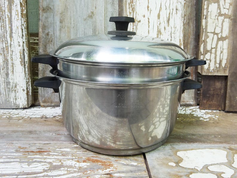 May include: A stainless steel stockpot with a matching lid and black plastic handles on both sides. The pot features a tiered design with a steamer insert, and the metal surface displays engraved text including the brand name Regal Ware.