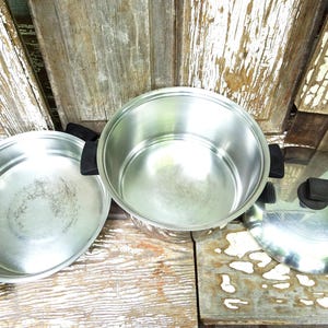 May include: A stainless steel cooking set consisting of a shallow pan, a deep pot, and a matching lid. Each piece features black plastic handles and a black knob on the lid. The interior surfaces are metallic silver with a polished finish.