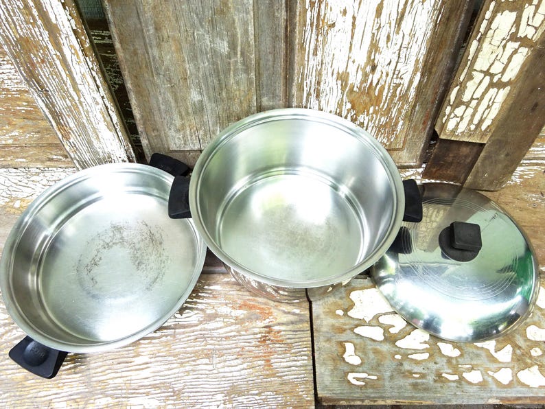 May include: A stainless steel cooking set consisting of a shallow pan, a deep pot, and a matching lid. Each piece features black plastic handles and a black knob on the lid. The interior surfaces are metallic silver with a polished finish.