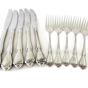 May include: Set of eight silver-toned flatware pieces, including five knives and three forks. The knives and forks have ornate handles with a floral design.
