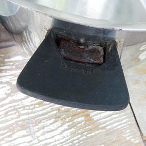May include: A silver-colored metal cooking pot featuring a black handle with visible rust accumulation at the connection point. The handle has the letters R and H embossed on its surface. The pot has a smooth, reflective metallic finish.
