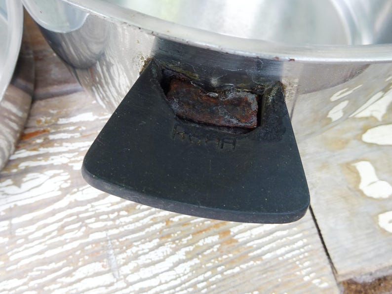 May include: A silver-colored metal cooking pot featuring a black handle with visible rust accumulation at the connection point. The handle has the letters R and H embossed on its surface. The pot has a smooth, reflective metallic finish.