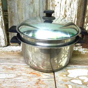 May include: A stainless steel stockpot with a matching lid and black heat-resistant handles on both sides. The pot features a polished metallic finish and a tiered design, with a secondary insert resting inside the main vessel under the lid.