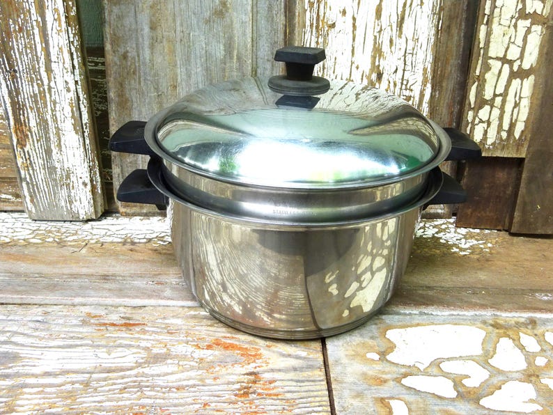 May include: A stainless steel stockpot with a matching lid and black heat-resistant handles on both sides. The pot features a polished metallic finish and a tiered design, with a secondary insert resting inside the main vessel under the lid.