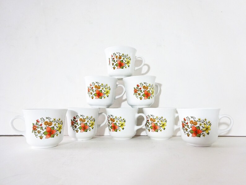 May include: Set of eight white ceramic mugs with a floral pattern in shades of orange, yellow, and green. The mugs are arranged in a pyramid shape.