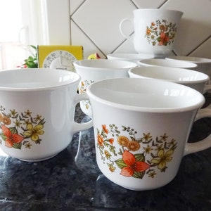 May include: Set of white ceramic mugs with floral designs in shades of orange, yellow, and green. The mugs have a rounded shape and a handle on the side.