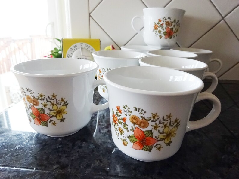 May include: Set of white ceramic mugs with floral designs in shades of orange, yellow, and green. The mugs have a rounded shape and a handle on the side.