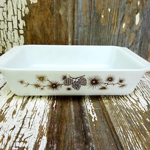 May include: A rectangular white glass baking dish with integrated side handles. The exterior features a brown graphic print of pine cones and needle-like branches centered on the long side of the vessel, providing a clear view of the interior.