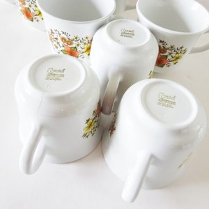 May include: Set of six white ceramic coffee mugs with a floral pattern in shades of brown, orange, and yellow. The mugs have a gold rim and the words "Corelle Impressions" are printed on the bottom.