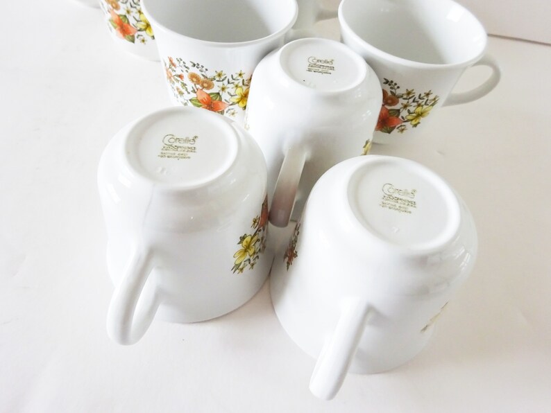 May include: Set of six white ceramic coffee mugs with a floral pattern in shades of brown, orange, and yellow. The mugs have a gold rim and the words "Corelle Impressions" are printed on the bottom.