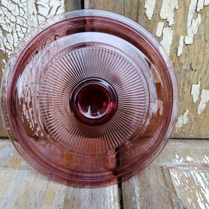 May include: A vintage glass cake plate with a pink hue and a raised, concentric circle design. The plate has a smooth, glossy finish.