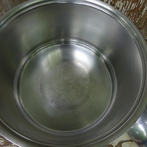 May include: A large, round, stainless steel cooking pot with a polished interior surface and a wide rim. The pot features two black side handles and is accompanied by a matching stainless steel lid with a circular design on its surface.