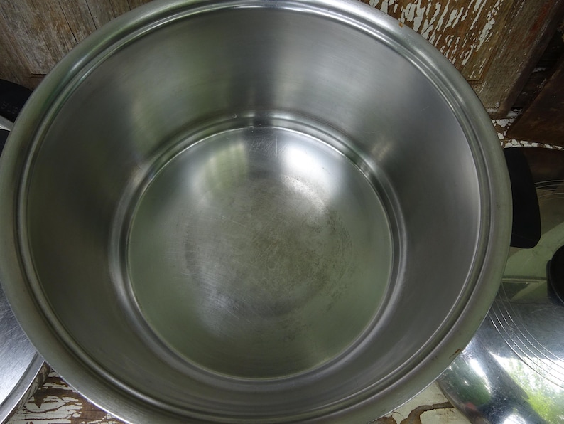 May include: A large, round, stainless steel cooking pot with a polished interior surface and a wide rim. The pot features two black side handles and is accompanied by a matching stainless steel lid with a circular design on its surface.