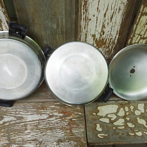 May include: Two round silver-colored metal pans with black plastic handles and one matching silver-colored metal lid. The pans and lid are shown from an overhead perspective, displaying their smooth, metallic surfaces and circular shapes.