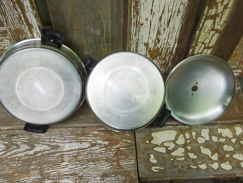 May include: Two round silver-colored metal pans with black plastic handles and one matching silver-colored metal lid. The pans and lid are shown from an overhead perspective, displaying their smooth, metallic surfaces and circular shapes.