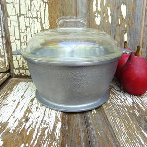 May include: A round, silver-colored metal cooking pot with a textured exterior surface, two small side handles, and a clear, domed glass lid featuring a subtle etched pattern. Two red pears are positioned next to the pot.