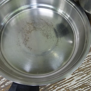 May include: A round stainless steel frying pan with a polished metallic interior surface showing light signs of use. The pan features a wide, flat base with slightly curved, raised sides and a black handle attachment visible at the bottom edge.