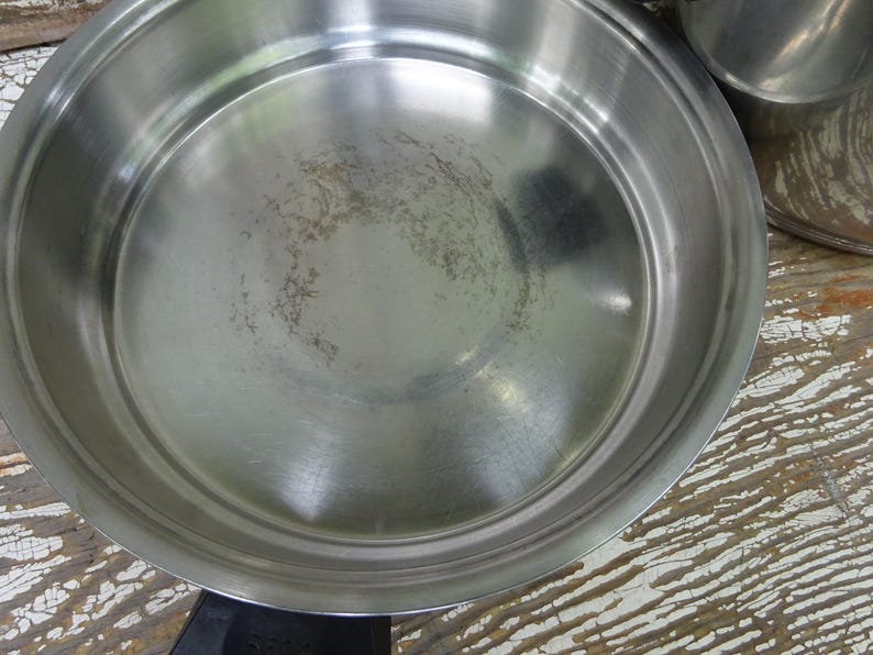 May include: A round stainless steel frying pan with a polished metallic interior surface showing light signs of use. The pan features a wide, flat base with slightly curved, raised sides and a black handle attachment visible at the bottom edge.