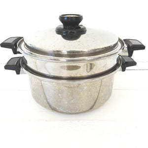 May include: A stainless steel two-tier steamer pot with a matching lid. The pot features black plastic handles on both sides of the base and the steamer insert, and a black knob on the center of the lid. The metal surface has a polished finish.