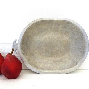 May include: An oval-shaped silver-toned metal bowl with a textured interior surface and two small side handles, positioned next to two red pears. The bowl features a matte finish and a wide rim, providing a simple and functional design for display.