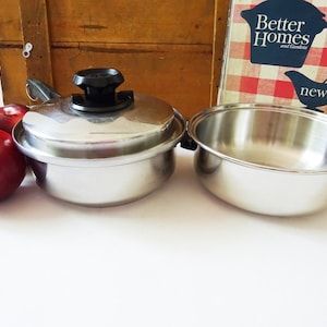 May include: A set of two stainless steel cookware pieces, a saucepan with a lid and a larger pot. The saucepan has a black knob on the lid. The cookware is on a white surface with red apples in the foreground.