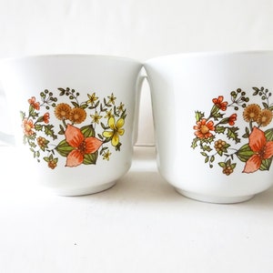 May include: Set of four white ceramic mugs with a floral design featuring orange, yellow, and green flowers. The mugs have a simple, rounded shape and a handle.