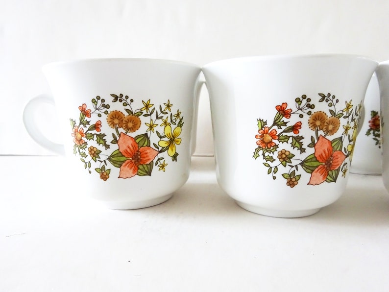 May include: Set of four white ceramic mugs with a floral design featuring orange, yellow, and green flowers. The mugs have a simple, rounded shape and a handle.