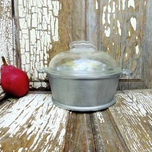 May include: A round, silver-colored metal cooking pot with a textured exterior surface and two small side handles. The pot is topped with a clear, domed glass lid featuring a decorative etched pattern. A single red pear sits to the left.