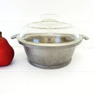 May include: A round, hammered metal serving dish with two side handles and a clear glass lid featuring an etched floral design. A single red pear is positioned to the left of the dish. The metal has a textured, matte gray finish.