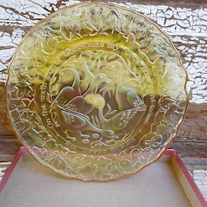 May include: Iridescent yellow glass plate with a raised design of geese and text from the Christmas carol "The Twelve Days of Christmas". The plate is in a pink and white box, set against a distressed wood background.