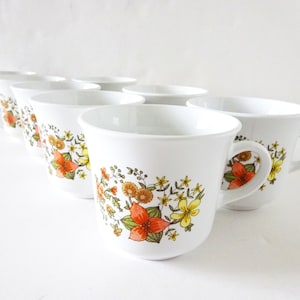 May include: Set of eight white ceramic teacups with a floral pattern in shades of orange, yellow, and green. The cups have a rounded shape and a handle.