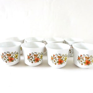 May include: Set of eight white ceramic mugs with a floral pattern in shades of orange, yellow, and green.