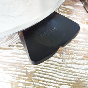 May include: A stainless steel pan featuring a black handle with the word RENA embossed on the top surface. The pan has a polished metallic finish and a wide, flat handle design that extends from the side of the circular cooking vessel.