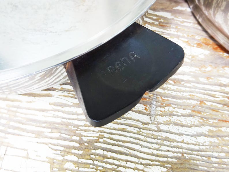 May include: A stainless steel pan featuring a black handle with the word RENA embossed on the top surface. The pan has a polished metallic finish and a wide, flat handle design that extends from the side of the circular cooking vessel.