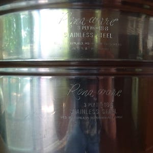 May include: A polished stainless steel cooking pot featuring the engraved text Rena-ware, 3 ply 18-8 stainless steel, and Made in U.S.A. The metallic surface is reflective, showing a cylindrical shape with a horizontal rim detail near the top edge.