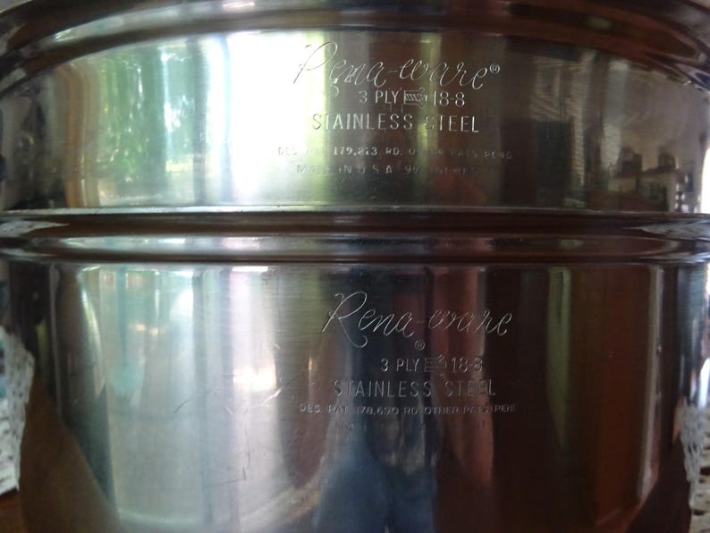 May include: A polished stainless steel cooking pot featuring the engraved text Rena-ware, 3 ply 18-8 stainless steel, and Made in U.S.A. The metallic surface is reflective, showing a cylindrical shape with a horizontal rim detail near the top edge.