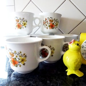 May include: Set of six white ceramic mugs with a floral pattern in shades of orange, yellow, and green. The mugs have a rounded shape and a handle on the right side.