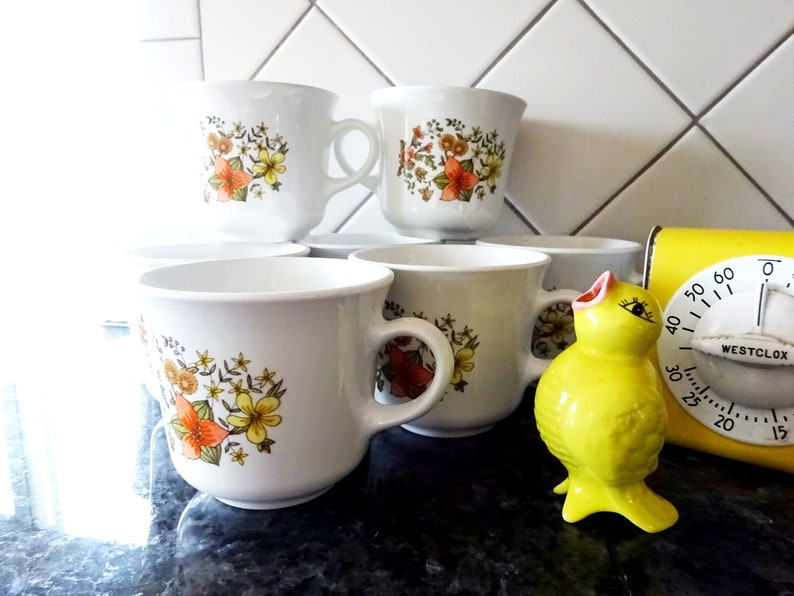 May include: Set of six white ceramic mugs with a floral pattern in shades of orange, yellow, and green. The mugs have a rounded shape and a handle on the right side.