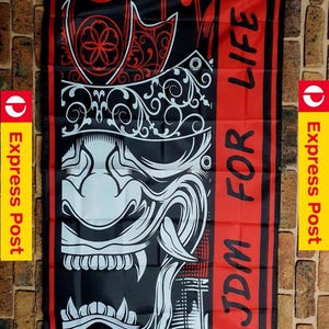 Könnte beinhalten: Eine rechteckige Flagge in Schwarz und Rot mit einer weißen Grafik einer stilisierten Samurai-Maske. Das Design umfasst komplizierte Schnörkelmuster und den fetten schwarzen Schriftzug JDM FOR LIFE, der vertikal auf einem roten Hintergrund gedruckt ist.