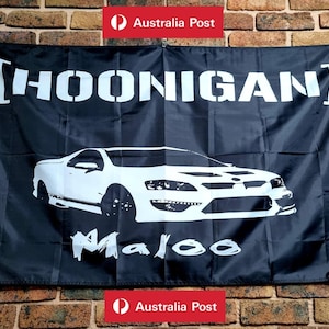 May include: A black rectangular flag featuring the white text HOONIGAN in brackets at the top, a white silhouette of a Maloo utility vehicle in the centre, and the word Maloo written in a stylised white font underneath the vehicle graphic.