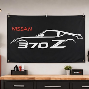 May include: A black rectangular wall banner featuring a white outline of a Nissan 370Z sports car in profile. The word NISSAN is printed in red above the car, and the text 370Z is printed in large white letters below the car silhouette.