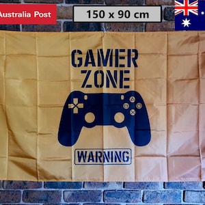 May include: A rectangular gold-coloured flag measuring 150 by 90 cm. The flag features the dark blue text GAMER ZONE at the top, a dark blue video game controller graphic in the centre, and the dark blue text WARNING inside a box at the bottom.