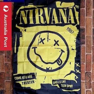 Puede incluir: Una bandera rectangular negra y amarilla con la palabra NIRVANA en letras grandes, el año 1987 y un gráfico central de cara sonriente con ojos en forma de X. El texto en la parte inferior dice COME AS U ARE THEATER y SMELLS LIKE TEEN SPRIT.