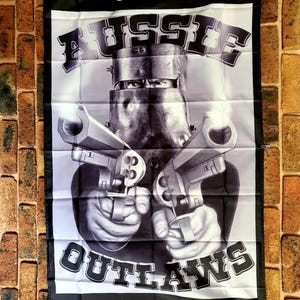 Peut inclure: Un drapeau rectangulaire en noir et blanc représentant une personne portant un casque en métal et tenant deux revolvers. Le texte indique "SUCH IS LIFE", "AUSSIE", "OUTLAWS" et les années "1855 - 1880" en caractères gras et bien visibles.