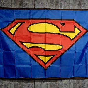 Può includere: Una bandiera rettangolare blu con l'iconico logo di Superman al centro. L'emblema è costituito da una grande forma a diamante rossa con un simbolo giallo a forma di S delineato in nero. La bandiera presenta un bordo marrone scuro e occhielli.