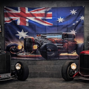 May include: A large rectangular wall flag featuring the Australian flag design overlaid with a graphic of a brown hot rod car. The flag is displayed on a grey brick wall between two vintage hot rod cars, one black and one red, parked in a garage.