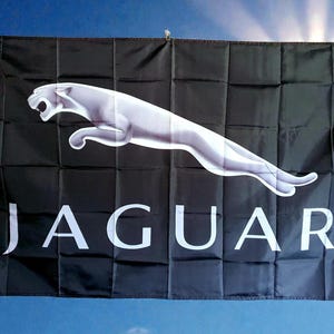 May include: A rectangular black flag featuring a large, silver-toned leaping jaguar graphic positioned above the white, capitalised text "JAGUAR". The fabric displays visible horizontal and vertical fold lines across the entire surface area.