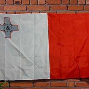 Peut inclure: Un drapeau rectangulaire de Malte composé de deux bandes verticales blanche et rouge. La section blanche sur la gauche présente l'emblème de la Croix de Georges dans le coin supérieur, bordé de rouge, avec une image centrale de Saint Georges.