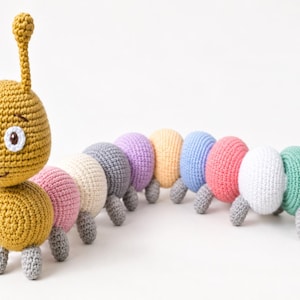 Puede incluir: Un juguete de oruga tejido a ganchillo con una cabeza de color amarillo mostaza, dos antenas, ojos blancos con pupilas marrones y un cuerpo largo segmentado en varios colores pastel, incluyendo rosa, crema, gris, lavanda, melocotón, azul, blanco, verde y lima.